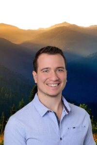 Aaron, a psilocybin facilitator at Vital Reset in Oregon, smiling against a forest and mountain backdrop. He supports veterans, first responders, and clients with trauma, anxiety, and depression through safe psilocybin treatment and holistic care.