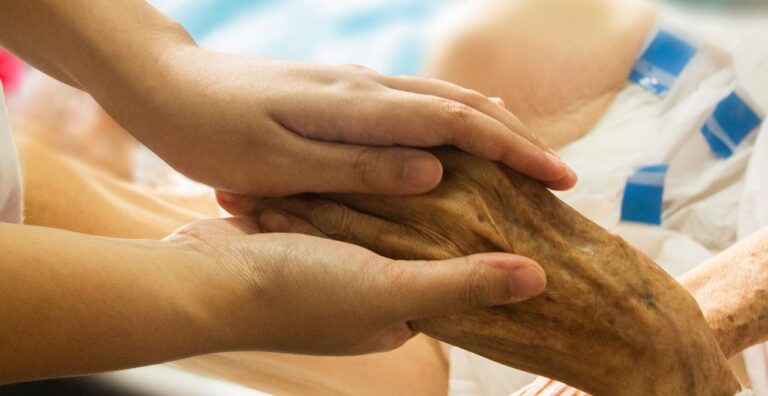 A younger hand gently holding the hand of an elderly person in a caregiving setting, symbolizing support and compassion for individuals living with Parkinson's Disease. Represents safe psilocybin treatment and the potential psilocybin benefits for aging populations in Oregon.