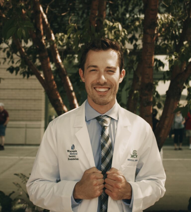 “Medical student Joe Hennesey, in white coat with Wisconsin Medical Society Foundation and MCW logos, smiling outdoors in front of trees.”