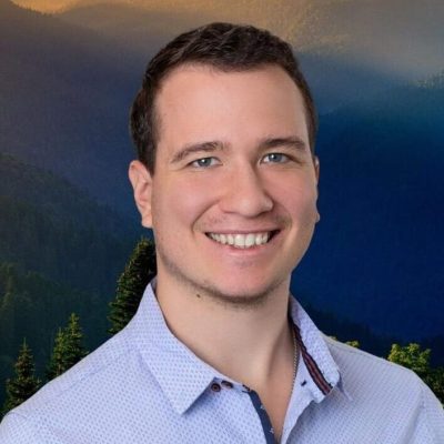 Aaron, a psilocybin facilitator at Vital Reset in Oregon, smiling against a forest and mountain backdrop. He supports veterans, first responders, and clients with trauma, anxiety, and depression through safe psilocybin treatment and holistic care.